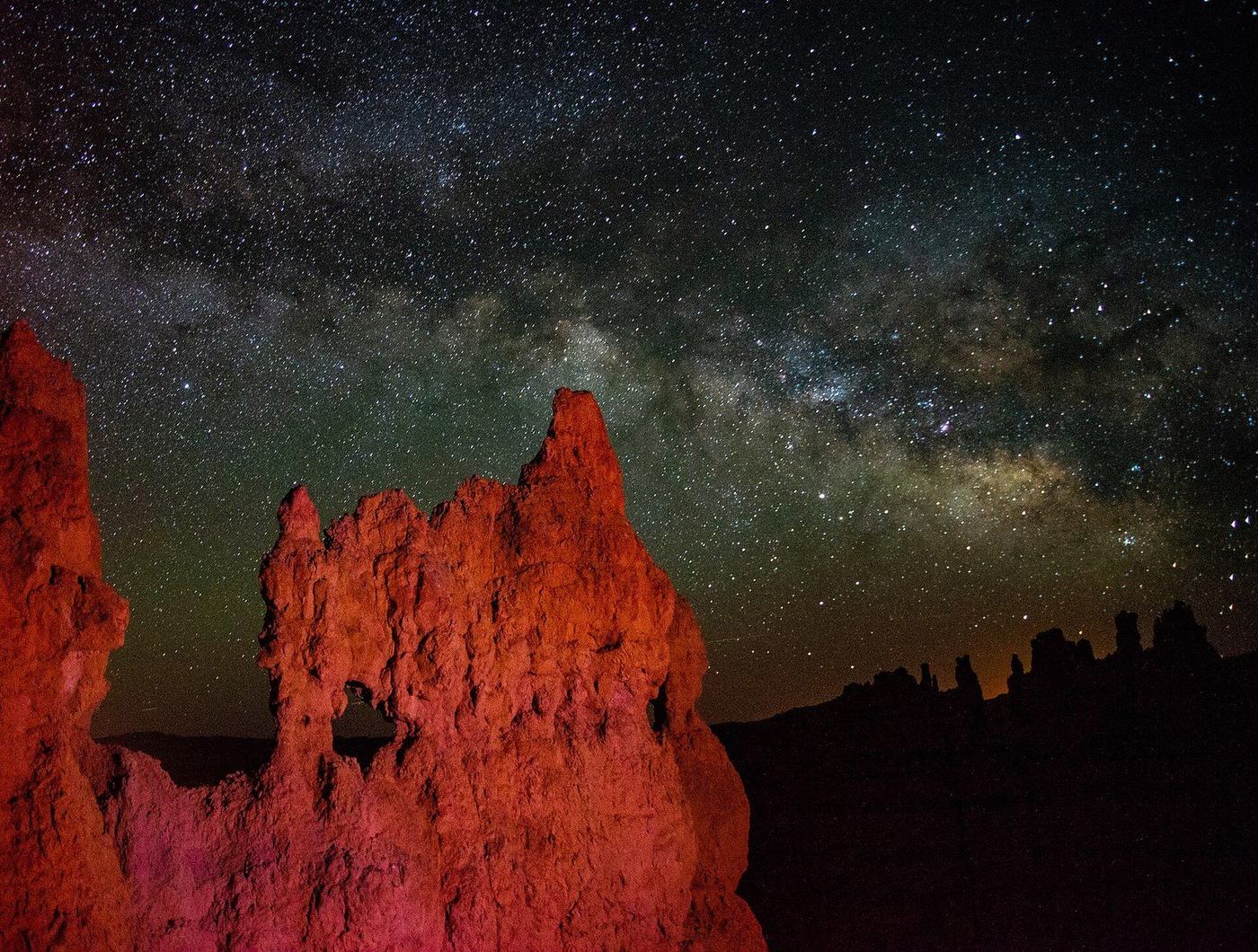 The Milky Way arching over red-lit hoodoos with a small window arch on the Navajo Trail in Bryce Canyon National Park