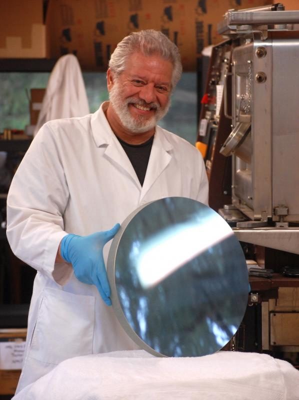 Carl Zambuto in a white lab coat and blue gloves, smiling and holding a large finished telescope mirror in his shop