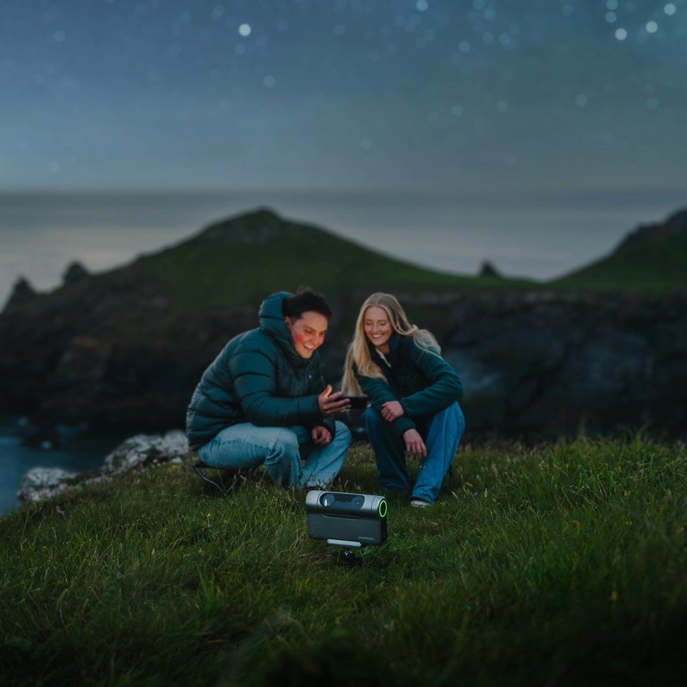 Two stargazers checking a phone beside a DwarfLab Dwarf 3 under a starry coastal sky