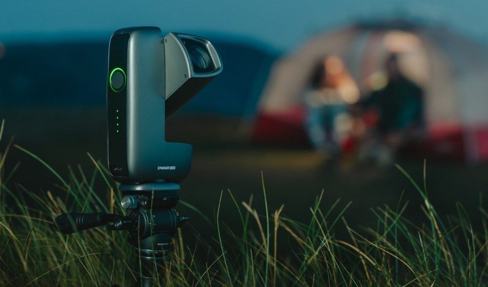 A DwarfLab smart telescope set up in tall grass beside a lit tent at a campsite, two people relaxing behind it &mdash; the take-it-anywhere smart-scope experience these two deliver