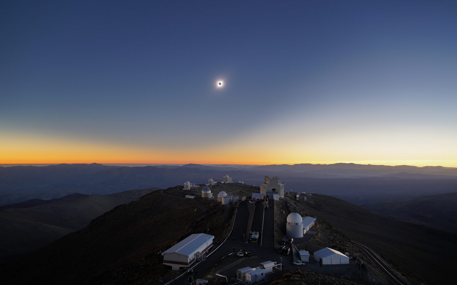 The eclipsed Sun hanging over the observatory domes of La Silla, Chile, with the horizon glowing sunset-orange in every direction