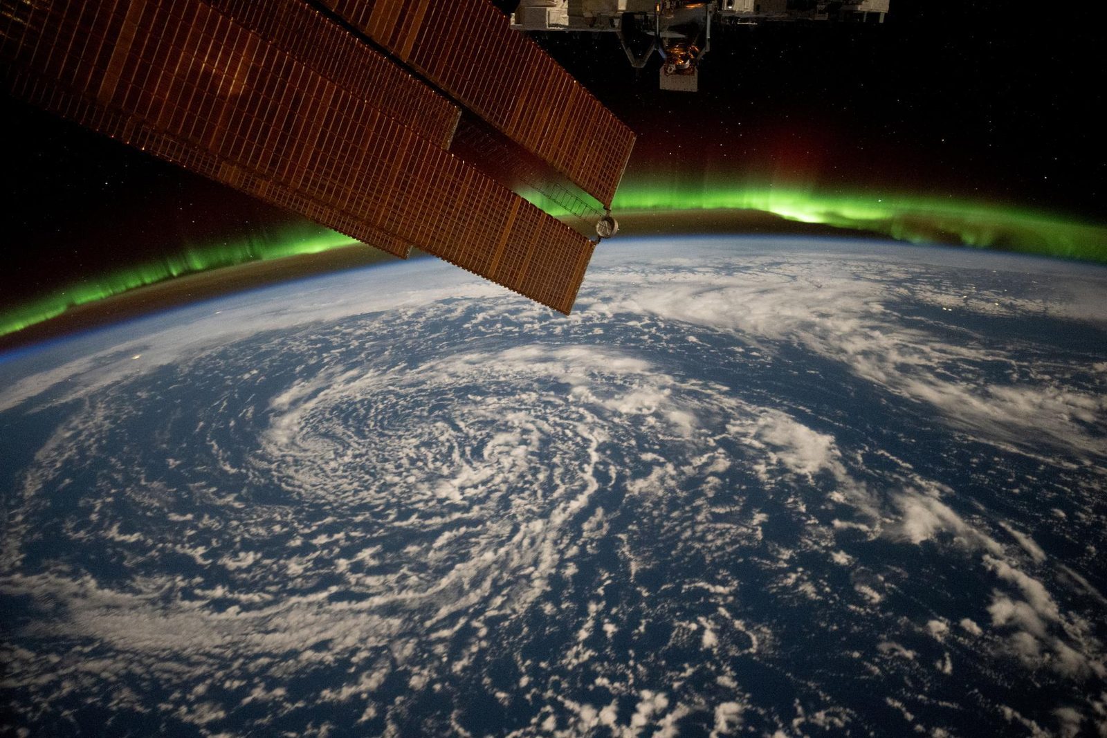 A green band of aurora borealis along Earth's horizon above swirling clouds over the Gulf of Alaska, photographed from the International Space Station with a solar array in the foreground