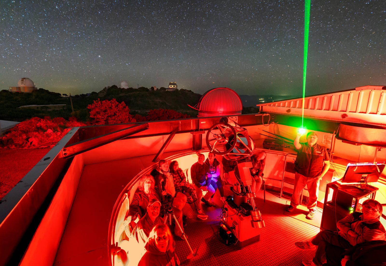 Visitors gathered under red light around a telescope in an open roll-off observatory during a public observing night, with domes and a star-filled sky behind them