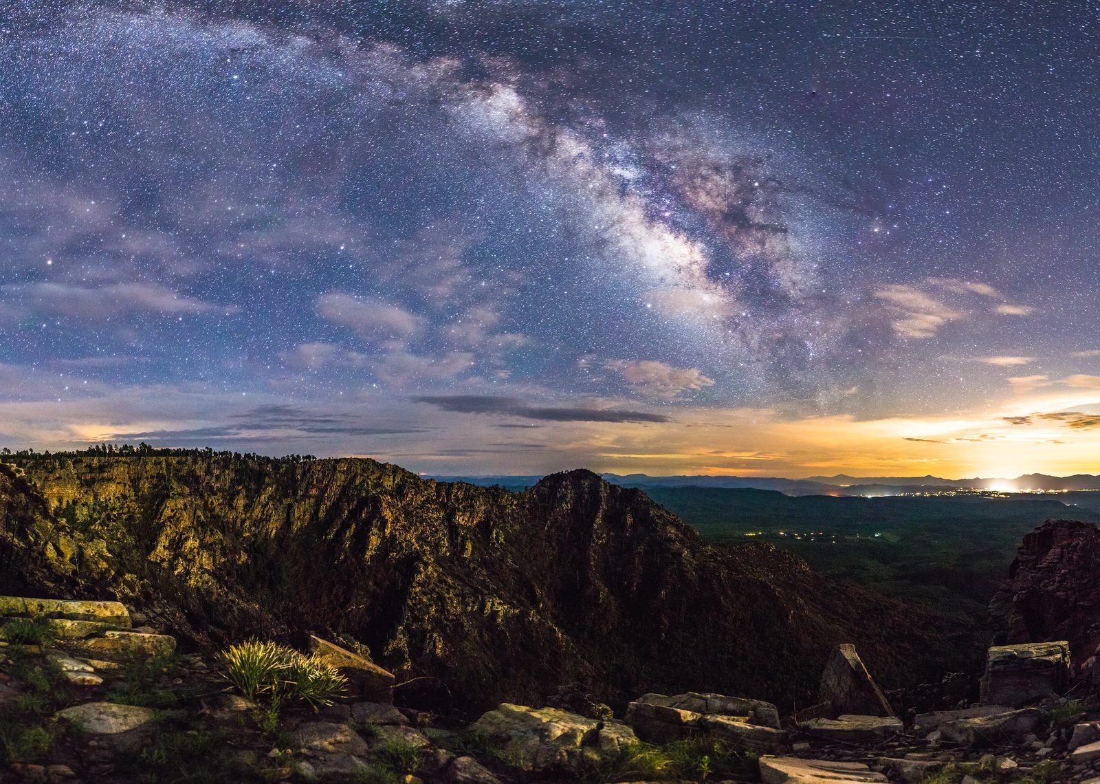 The Milky Way's core arcing over the rocky edge of the Mogollon Rim at night, with the lights of Payson on the horizon