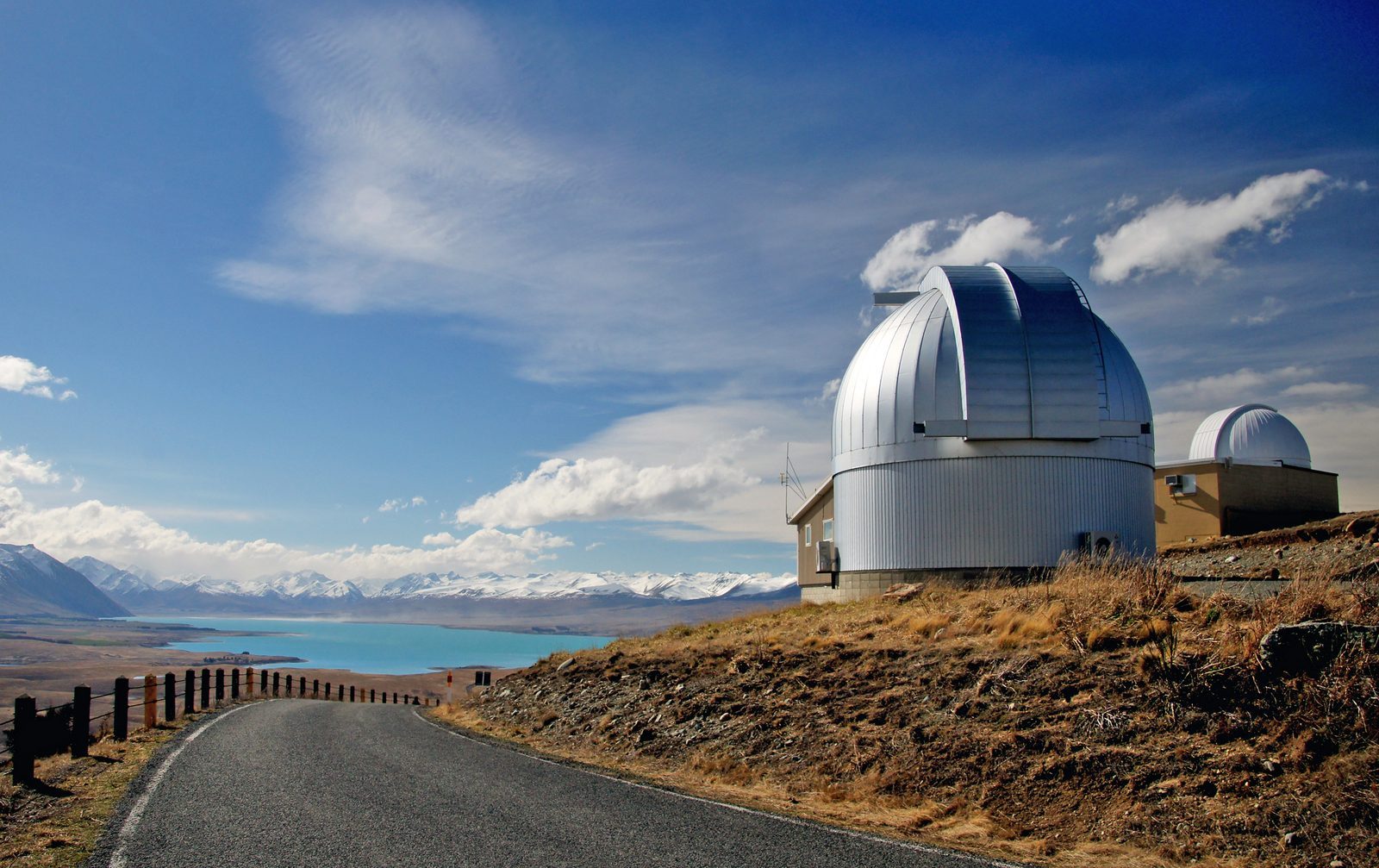 A silver telescope dome of the University of Canterbury Mount John Observatory above turquoise Lake Tekapo, with snow-capped mountains beyond