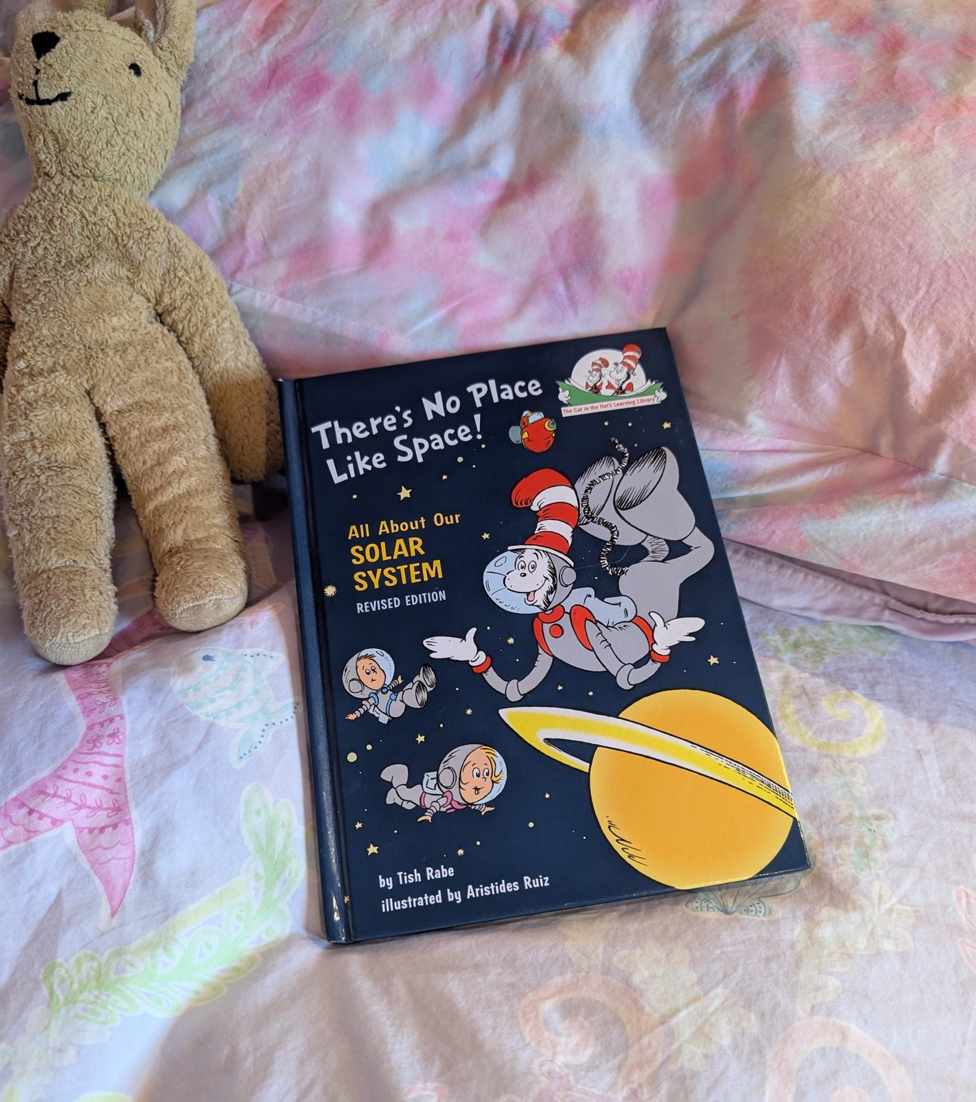A hardcover copy of There&rsquo;s No Place Like Space! All About Our Solar System, revised edition, propped on a child&rsquo;s bed beside a well-worn stuffed rabbit