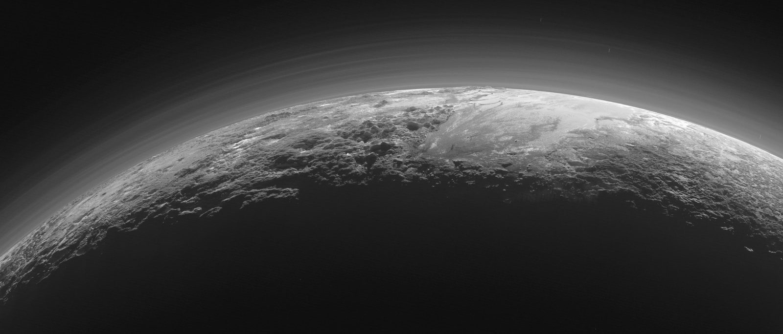 Pluto&rsquo;s ice mountains, frozen nitrogen plains and layered blue atmospheric haze, backlit near sunset &mdash; captured by NASA&rsquo;s New Horizons in July 2015