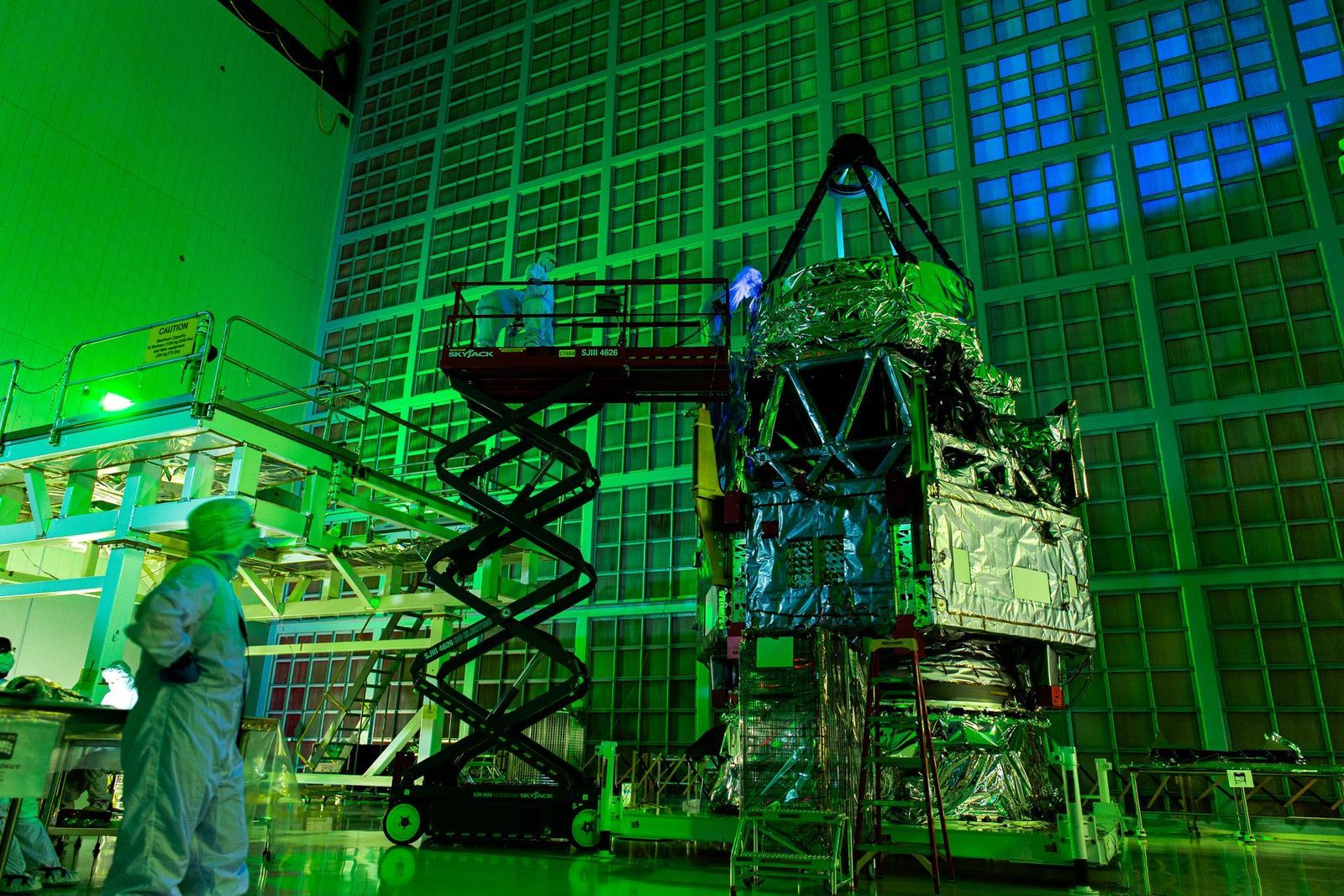 Roman Space Telescope team members on a scissor lift inspecting the observatory&rsquo;s 2.4-meter primary mirror with ultraviolet light in a darkened Goddard clean room, a long exposure washing the room green
