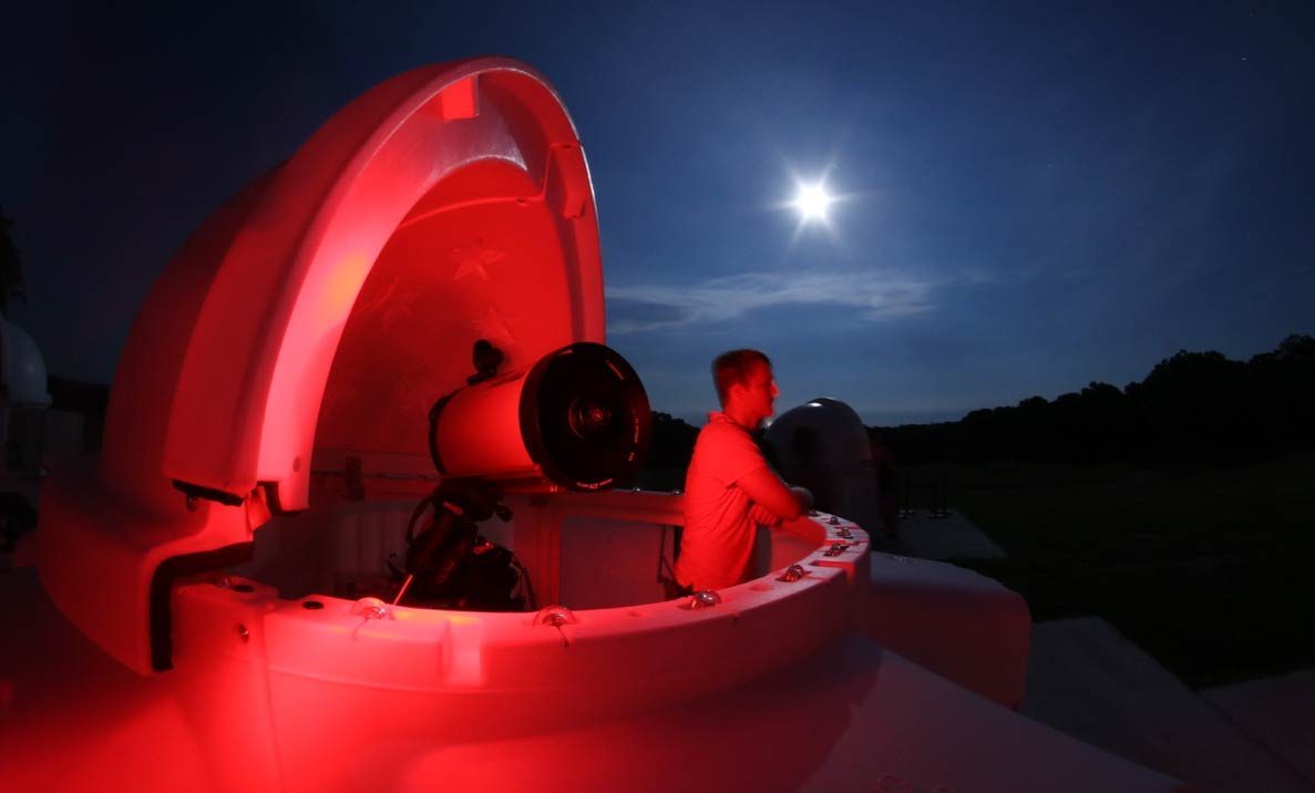 A SkyShed POD dome observatory open at dusk, its interior lit red, an observer at the eyepiece under a bright Moon