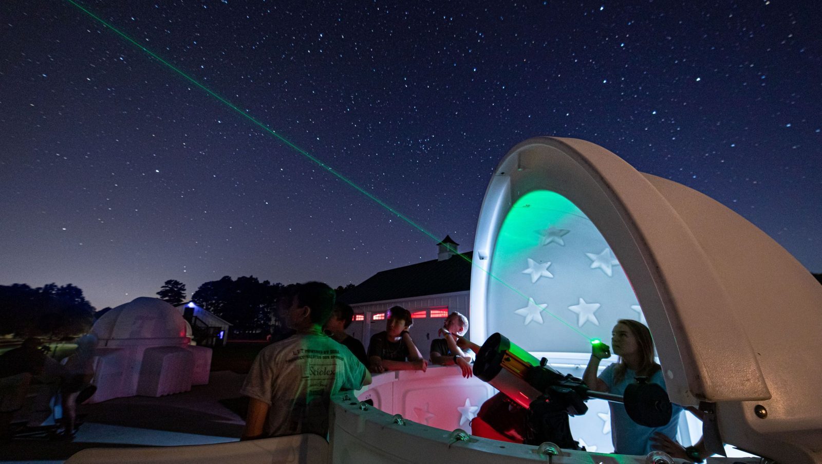 A group at a SkyShed POD observatory at night, one pointing a green laser at a star-filled sky beside a telescope, with a second POD in the background