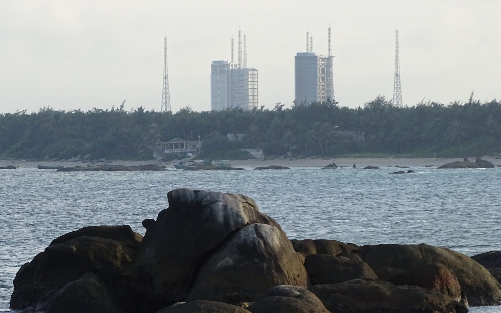 The launch towers of the Wenchang Space Launch Site on the horizon, seen across the water from the boulders of Qishui Bay, Hainan
