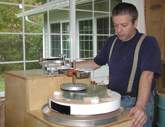 Carl Zambuto in suspenders standing at an early mirror-figuring machine in his shop, a mirror turning on the table beneath the polishing arm