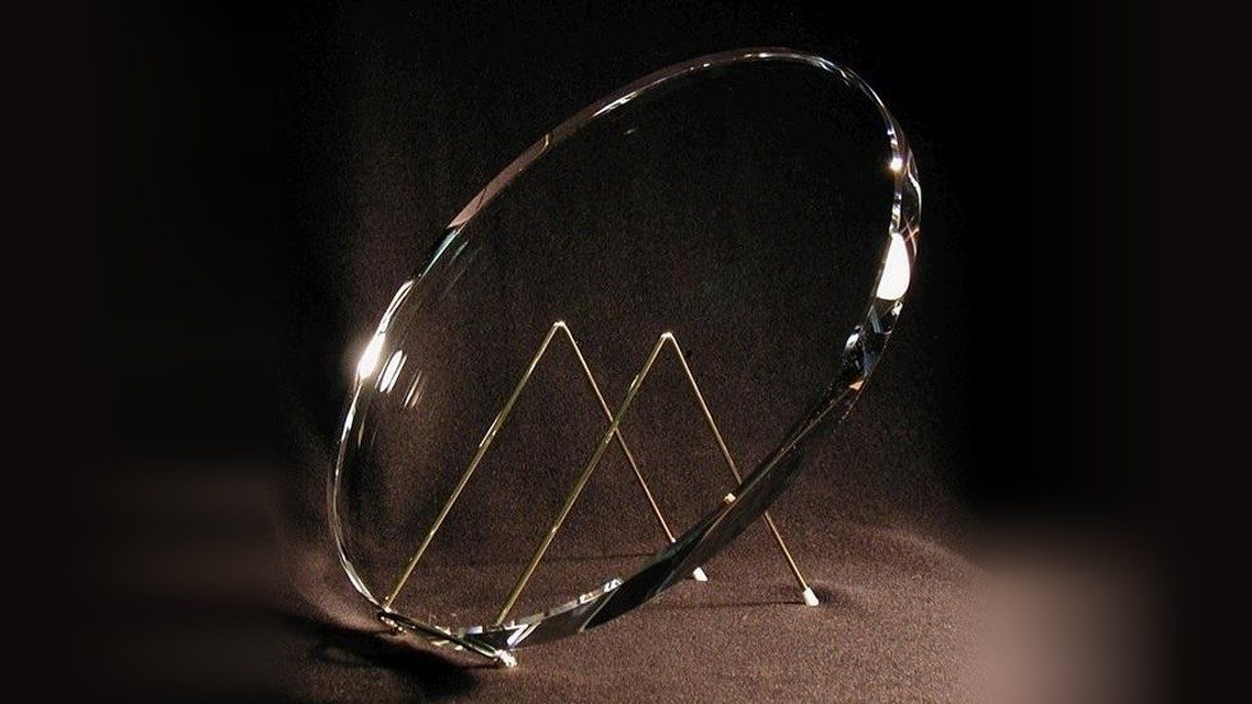 A finished Zambuto telescope mirror standing on edge on a wire stand against black cloth, its bevel catching the light