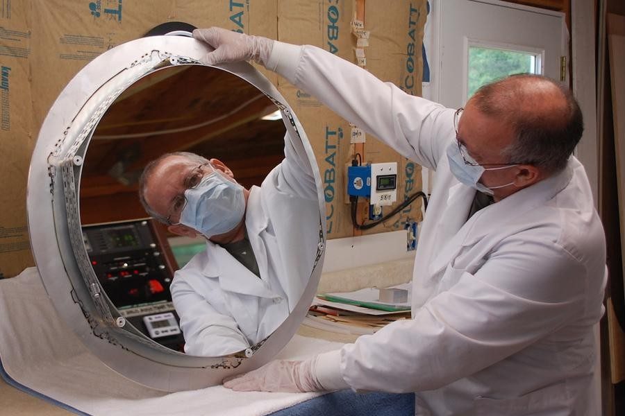 A technician in a lab coat, mask and gloves holding up a freshly coated 24-inch Zambuto mirror in its ring, his reflection looking back from the glass