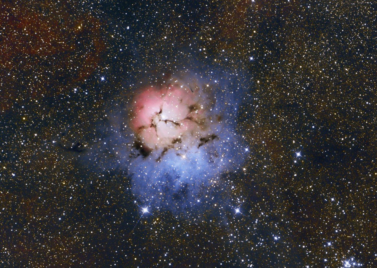 The Trifid Nebula, a pink cloud split by dark dust lanes beside a blue glow, in a dense starfield, photographed through a 12.5-inch Zambuto mirror